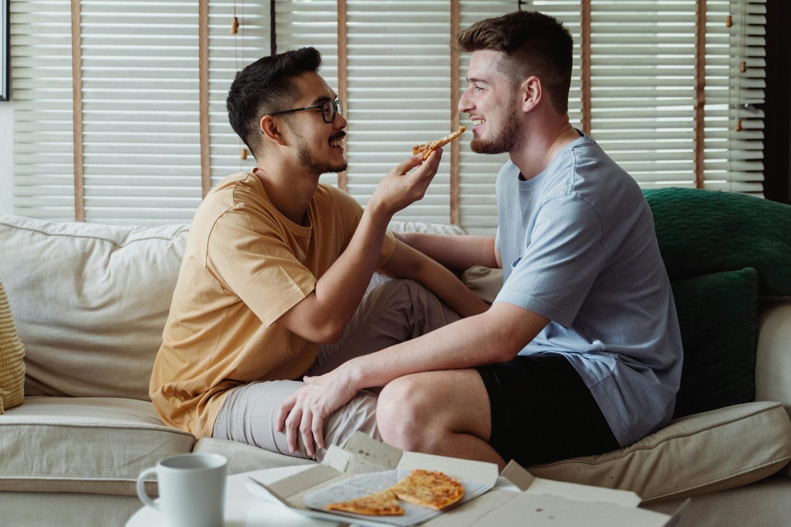Happy interracial LGBT couple enjoying each other's company and pizza on a cozy sofa indoors.